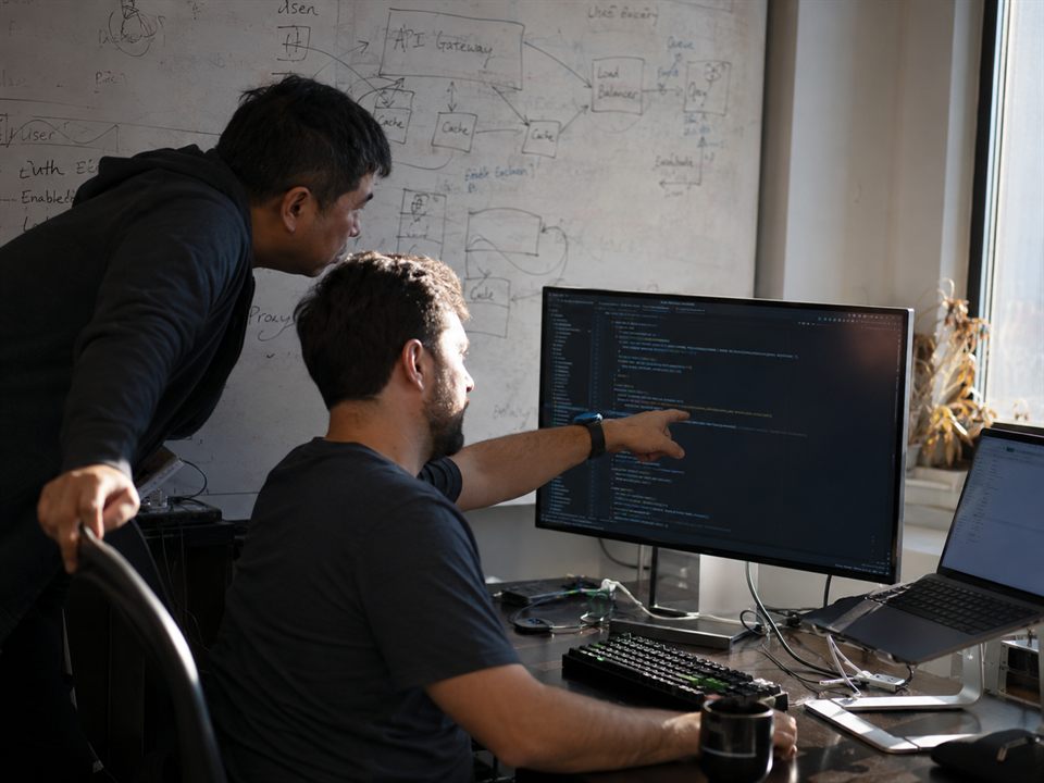 Two engineers debugging code together at one desk, with a system architecture diagram on the whiteboard behind them