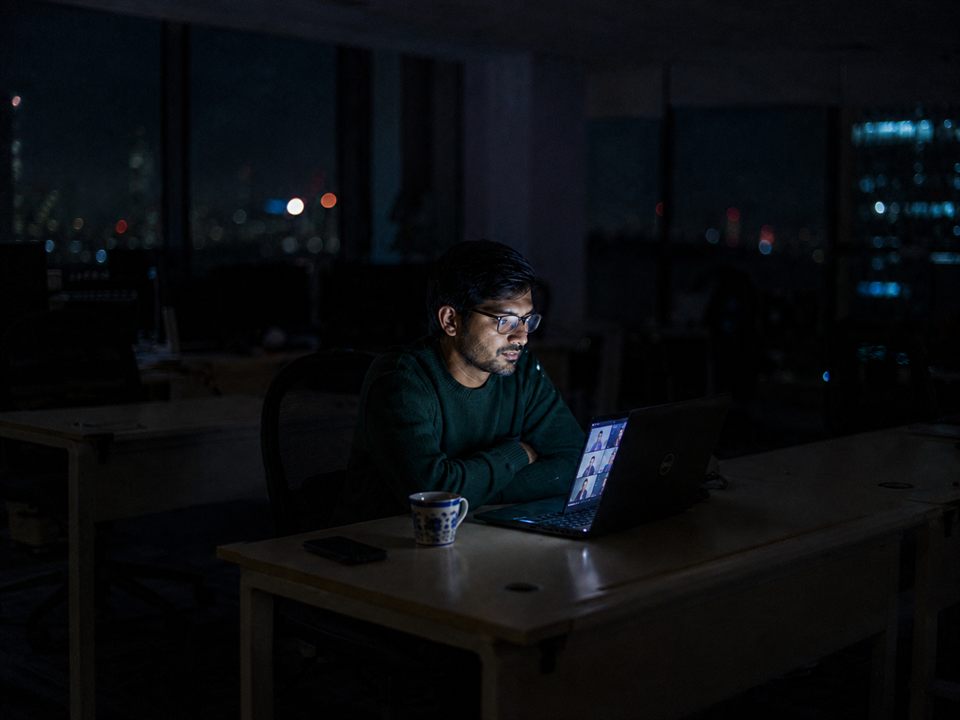 An engineer on a late-night video call with colleagues in another timezone, city lights behind him